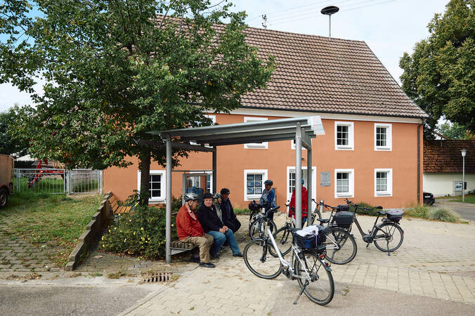 Vor dem Rathaus Sumpfohren sitzen drei Personen auf einer Bank unter einem Baum, während mehrere Fahrräder daneben abgestellt sind.