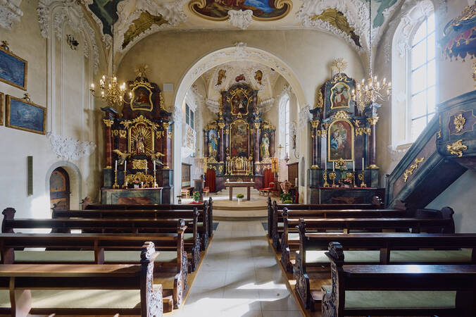 Ein barocker Kirchenraum mit prächtigen Deckenmalereien, vergoldeten Altären und Holzbänken, erhellt von einem Kronleuchter.
