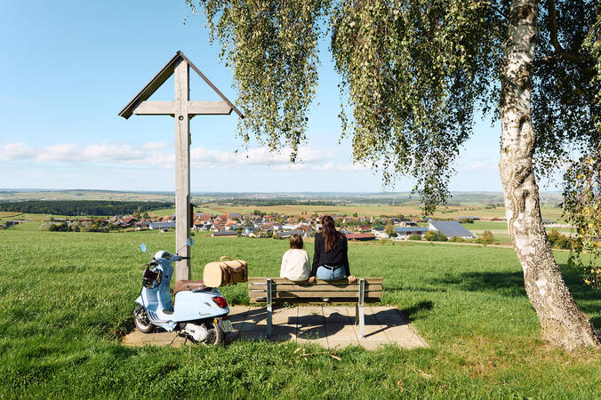 Zwei Personen sitzen auf einer Bank neben einem Motorroller und einem Kreuz, blicken auf eine ländliche Landschaft mit einem Dorf im Hintergrund.