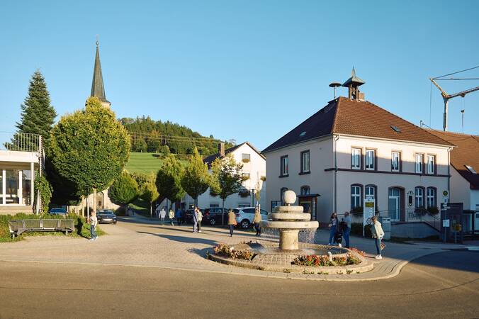 Die Dorfmitte von Fürstenberg mit einem Brunnen in der Mitte, umgeben von Bäumen, einer Kirche im Hintergrund und mehreren Menschen, die spazieren gehen.