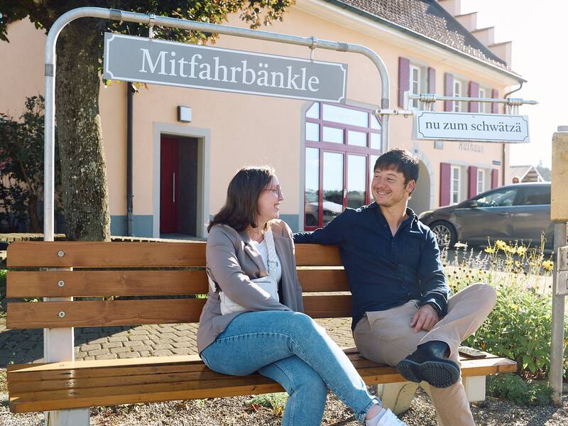 Zwei Personen sitzen entspannt auf einer Holzbank unter einem Schild mit der Aufschrift "Mitfahrbänkle".