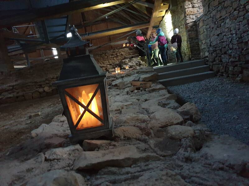 Im Bild ist ein beleuchteter Raum mit einer alten Steinmauer und einer großen Laterne im Vordergrund zu sehen, während im Hintergrund mehrere Personen mit Helmen eine Treppe hinaufsteigen.