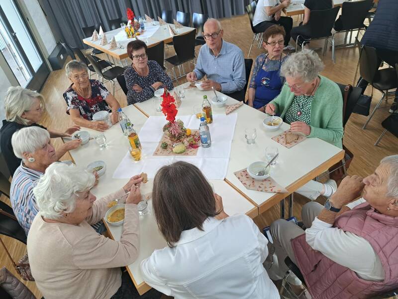 Eine Gruppe älterer Menschen sitzt um einen rechteckigen Tisch, isst Suppe und unterhält sich in einem Raum mit gedeckten Tischen.