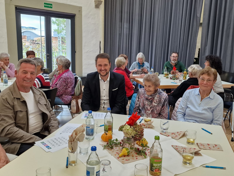 Eine Gruppe von Menschen sitzt fröhlich an einem Tisch in einem Raum mit großen Fenstern und Vorhängen, wobei der Tisch mit Blumen und Getränken dekoriert ist. Dabei wird Bingo im Rahmen der Veranstaltungsreihe der Nachbarschaftshilfe Hüfingen gespielt.