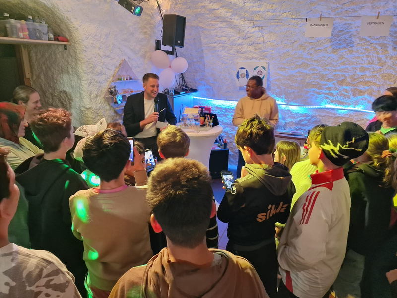 Bürgermeister Patrick Haas spricht in einem Raum vor einer Gruppe von Kindern und Jugendlichen mit Mikrofon, während der neue Leiter des Jugendhauses Jackson Agbonkhese  neben ihm steht.