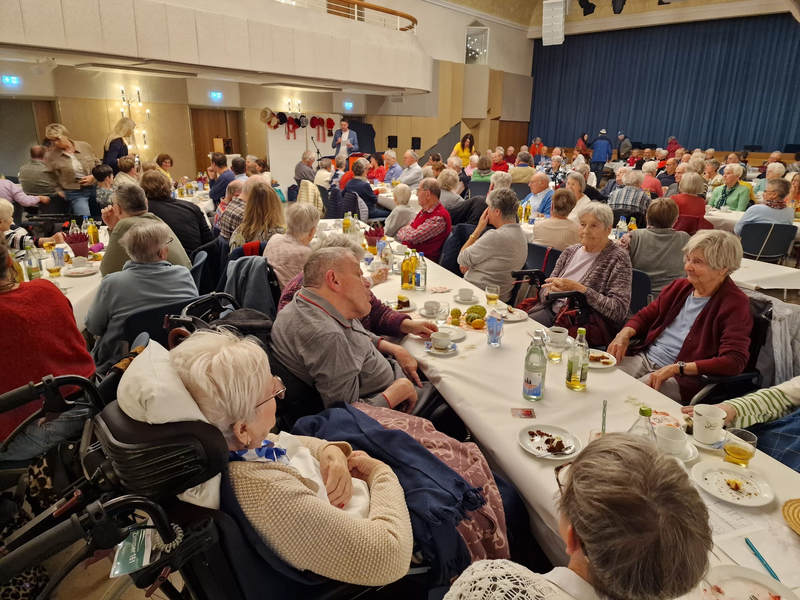 Die Stadthalle Hüfingen ist gefüllt mit älteren Menschen, die an langen, gedeckten Tischen sitzen, miteinander plaudern und Snacks genießen.