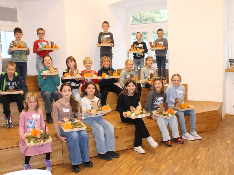 Eine Gruppe von Kindern sitzt auf Holzstufen in der Kinder- und Jugendbibliothek und hält dekorierte Tabletts mit herbstlichen Motiven in den Händen.