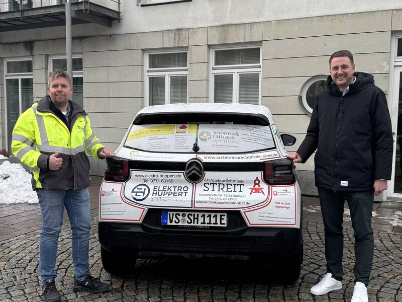 Bauamtsleiter Thilo Mayer und Bürgermeister Patrick Haas stehen neben dem neuen städtischen Elektrofahrzeug, das mit den Logos der Sponsoren beklebt ist.