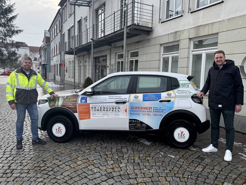 Bauamtsleiter Thilo Mayer und Bürgermeister Patrick Haas stehen neben dem neuen städtischen Elektrofahrzeug, das mit den Logos der Sponsoren beklebt ist.