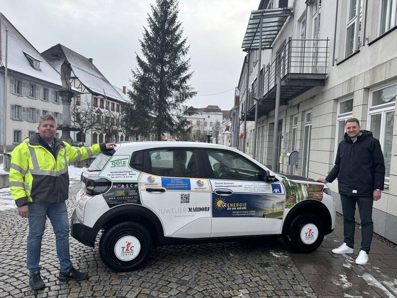 Bauamtsleiter Thilo Mayer und Bürgermeister Patrick Haas stehen neben dem neuen städtischen Elektrofahrzeug, das mit den Logos der Sponsoren beklebt ist.