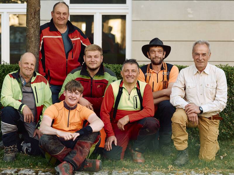 Eine Gruppe von 7 Forstmitarbeitern sitzt lächelnd in Arbeitskleidung vor dem Rathaus.