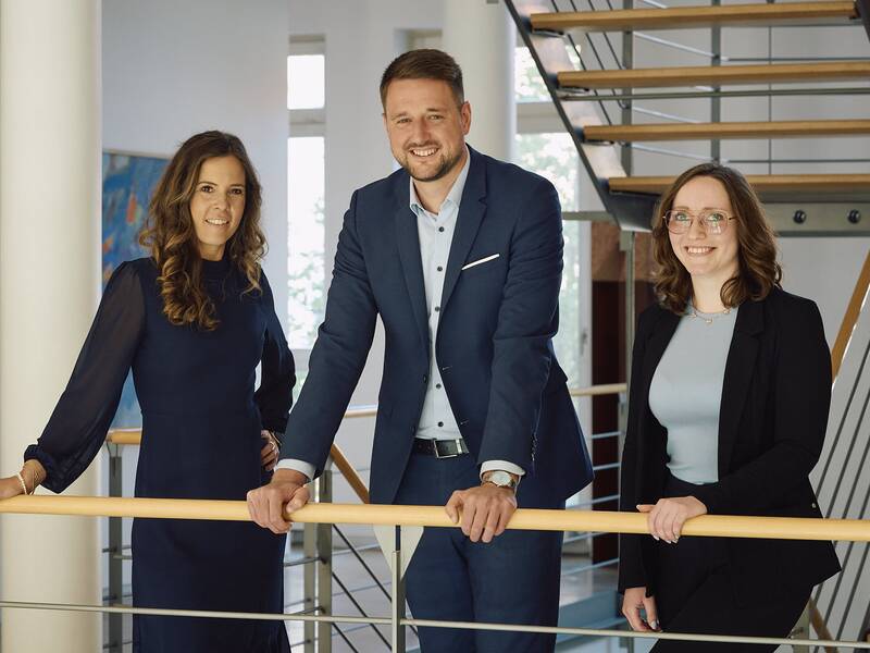 Die Mitarbeiter des Referates Bürgermeister & Gemeinderat, Frau Kirsten Frade Babau, Herr Bürgermeister Patrick Haas und Frau Stefanie Willmann (von links nach rechts) stehen lächelnd im modernen Rathausgebäude an einem Geländer.