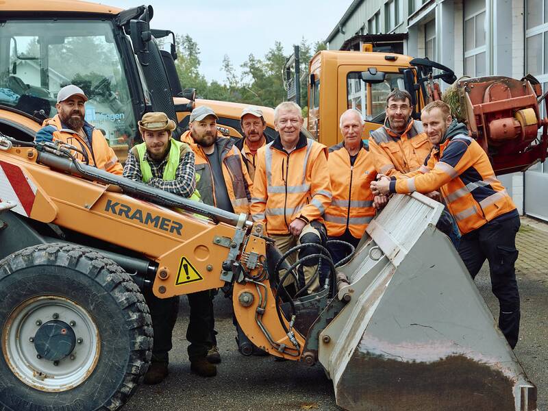 Die Mitarbeiter des Bauhofes stehen lächelnd vor dem Fuhrpark des Bauhofes.