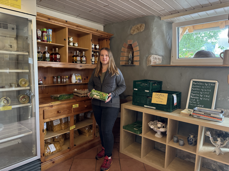 Larissa Baur steht im eigenen Hofladen des Breite-Halde-Hof in Hausen vor Wald, der mit Holzmöbeln und Regalen mit verschiedenen Lebensmitteln wie Saucen, Eiern und frischem Gemüse bestückt ist.