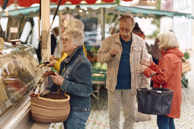 Mehrere Menschen stehen fröhlich an einer Markttheke des Hüfinger Wochenmarktes, eine Person davon mit einem Korb in der Hand, während sie miteinander sprechen.