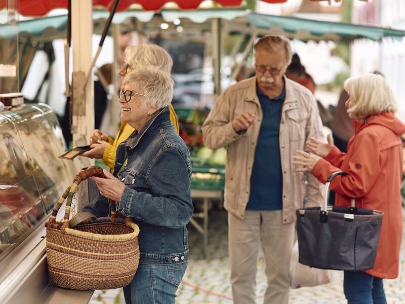 Mehrere Menschen stehen fröhlich an einer Markttheke des Hüfinger Wochenmarktes, eine Person davon mit einem Korb in der Hand, während sie miteinander sprechen.