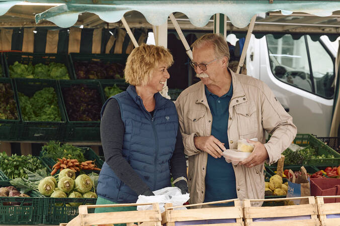 Eine Frau und ein Mann unterhalten sich fröhlich an einem Marktstand des Hüfinger Wochenmarktes mit frischem Gemüse im Hintergrund.