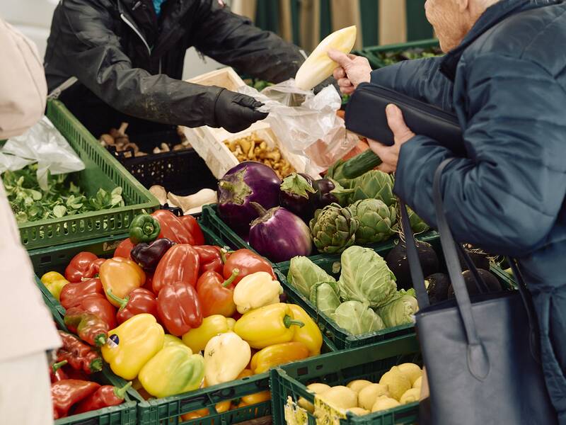 Eine Person auf einem Marktstand des Hüfinger Wochenmarktes greift nach frischem Gemüse wie Paprika, Auberginen und Artischocken.